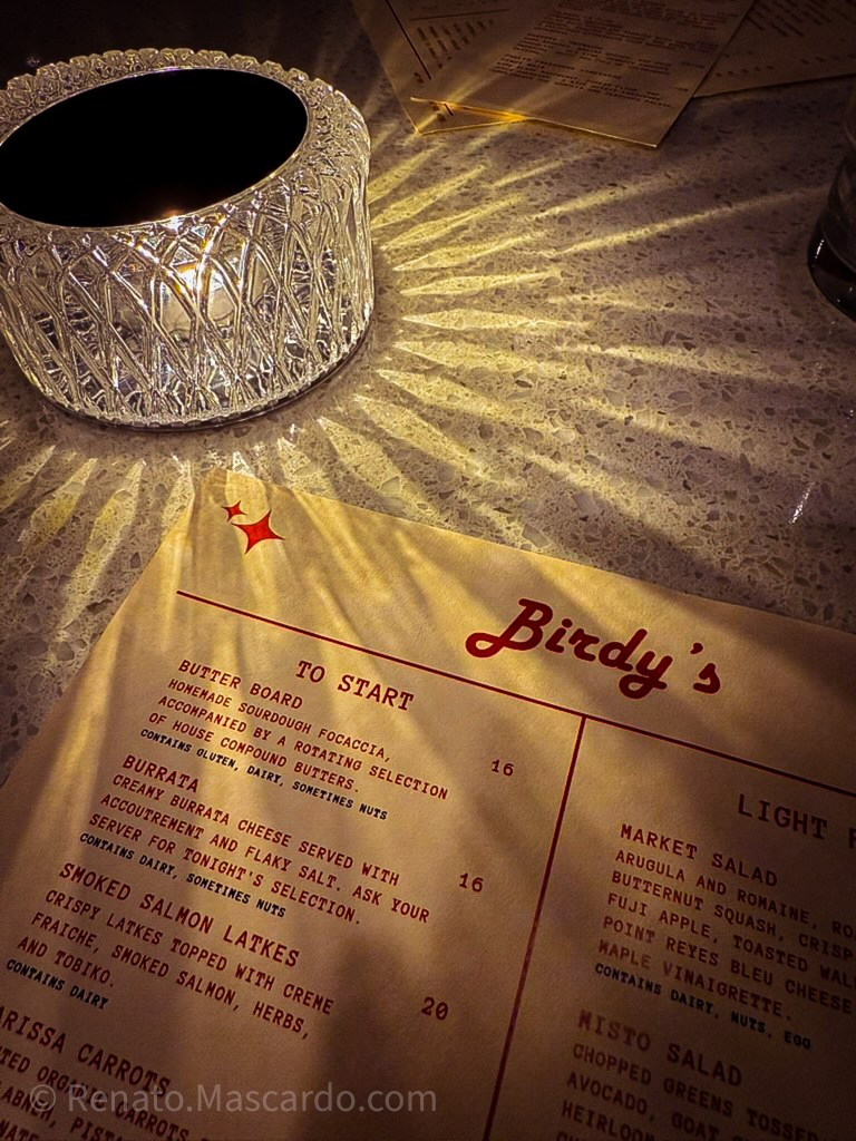 A close-up of a menu at Birdy's restaurant illuminated by candlelight, showcasing various dishes under the 'To Start' and 'Light Fare' sections.
