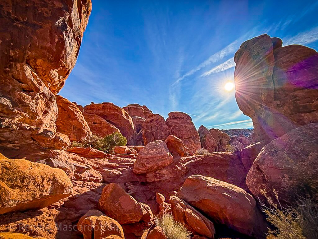 Sunlight streaming through rock formations in Arches National Park, highlighting red sandstone against a clear blue sky.