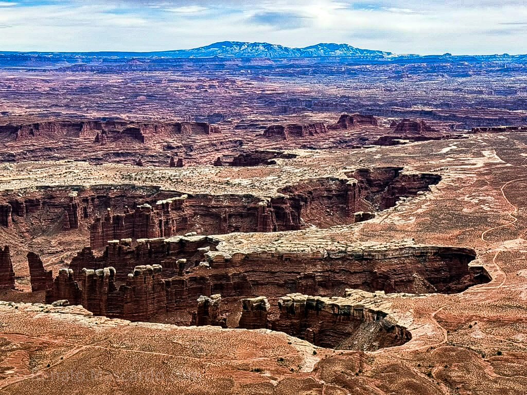 Panoramic view of the vast, rugged landscape of Canyonlands National Park, showcasing deep canyons, rock formations, and distant mountains under a cloudy sky.