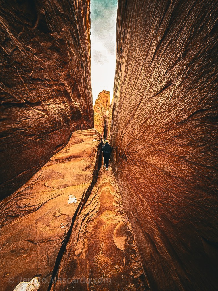 A person walking through a narrow sandstone canyon with steep walls, highlighting the texture and colors of the rock formations.