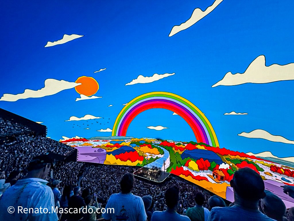 A vibrant and colorful digital landscape featuring a rainbow and abstract scenery, displayed prominently at a live concert venue, with an engaged audience visible in the foreground.