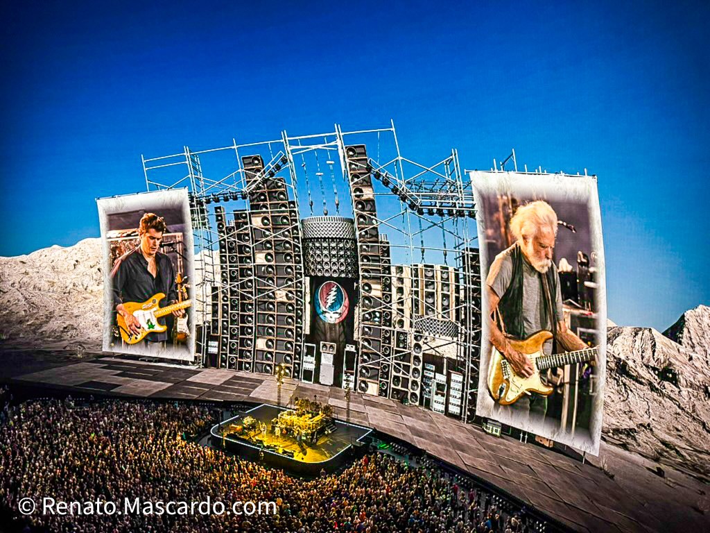 A large outdoor concert scene featuring a crowd in front of a grand stage with elaborate sound equipment, two images of musicians performing displayed on large screens.