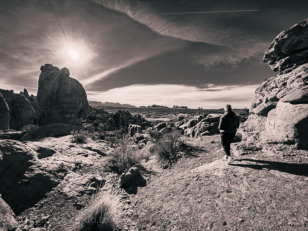 A person standing on rocky terrain in a desert landscape, with the sun casting a bright light in the background.