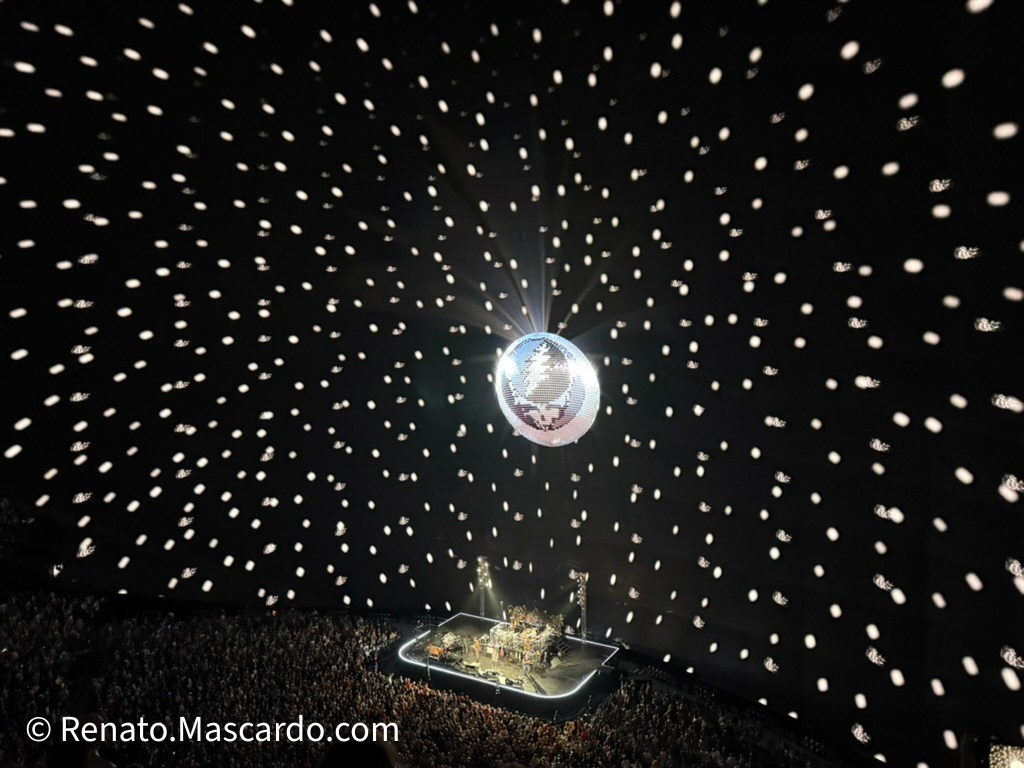 Aerial view of a concert performance inside The Sphere, featuring a disco ball with light projections creating a starry effect above a large audience.