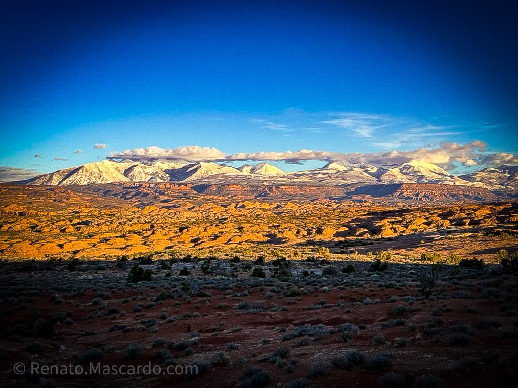 A panoramic view of the Utah landscape at sunset, featuring snow-capped mountains in the distance and a terrain of golden rock formations and sparse vegetation.