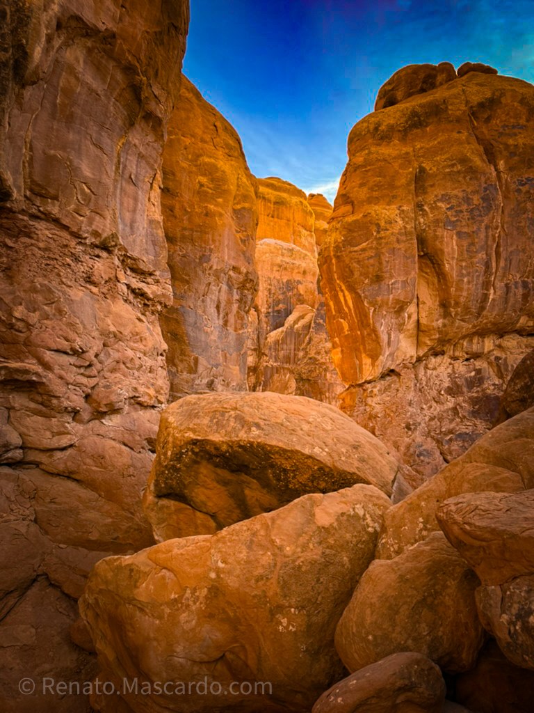 A narrow canyon with towering reddish rock formations and a vibrant blue sky above.