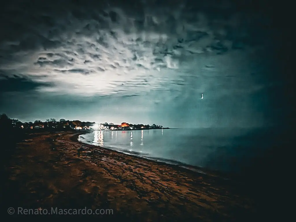 A serene night landscape featuring a beach with soft waves lapping at the shore, illuminated by distant lights from a quaint town and a cloudy sky overhead.