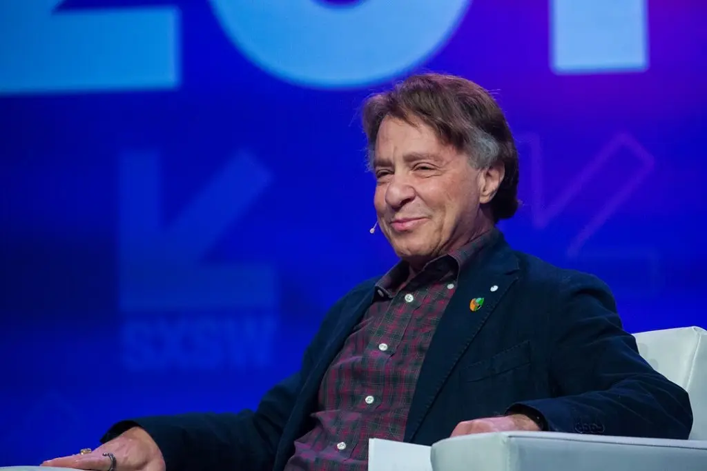 Ray Kurzweil speaking at a technology conference, smiling and wearing a dark blazer over a plaid shirt.