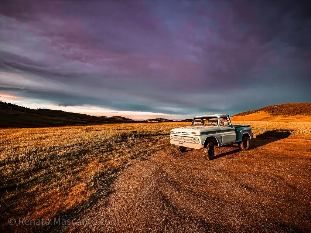 An old vintage pickup truck parked on a dirt road in a scenic landscape with grasslands and rolling hills under a colorful twilight sky.