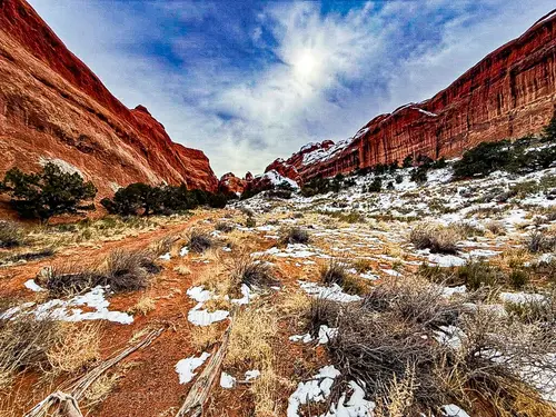 A scenic view of red rock formations and a snow-dusted desert landscape in Utah, under a cloudy sky.