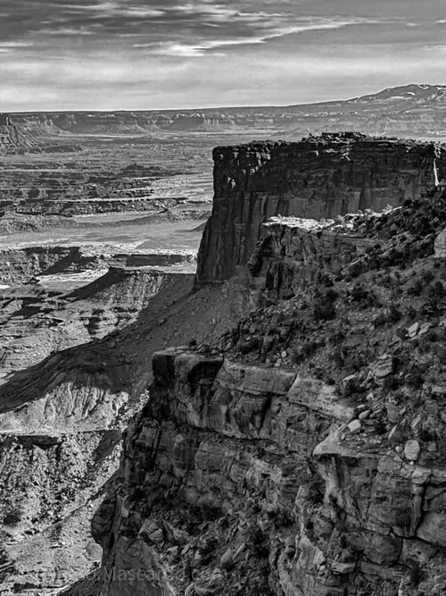 Black and white photograph of a dramatic cliff overlooking a vast canyon landscape, highlighting the rugged rock formations and distant mountains under a cloudy sky.