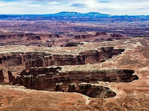 Panoramic view of the vast, rugged landscape of Canyonlands National Park, showcasing deep canyons, rock formations, and distant mountains under a cloudy sky.