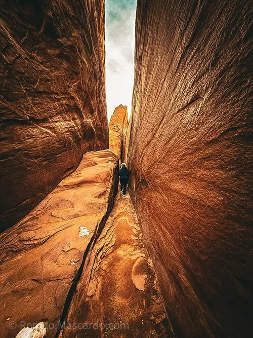 A person walking through a narrow sandstone canyon with steep walls, highlighting the texture and colors of the rock formations.