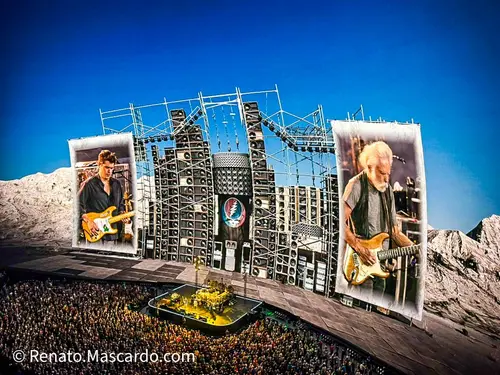 A large outdoor concert scene featuring a crowd in front of a grand stage with elaborate sound equipment, two images of musicians performing displayed on large screens.
