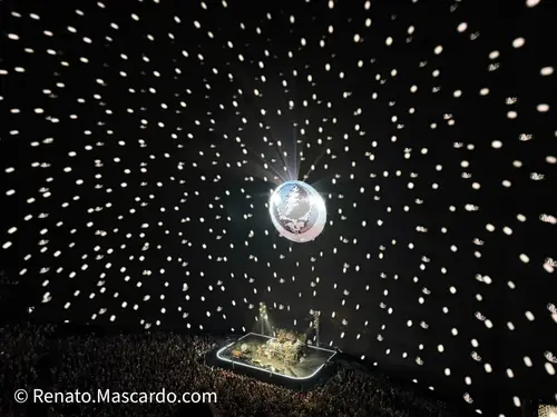 Aerial view of a concert performance inside The Sphere, featuring a disco ball with light projections creating a starry effect above a large audience.